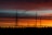 A series of electricity pylons and power lines are silhouetted against a vibrant sunset. The sky is painted with layers of orange, red, and blue hues, creating a dramatic backdrop. The landscape at the bottom shows trees and smaller structures in shadow, emphasizing the height and scale of the pylons.