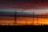 A series of electricity pylons and power lines are silhouetted against a vibrant sunset. The sky is painted with layers of orange, red, and blue hues, creating a dramatic backdrop. The landscape at the bottom shows trees and smaller structures in shadow, emphasizing the height and scale of the pylons.