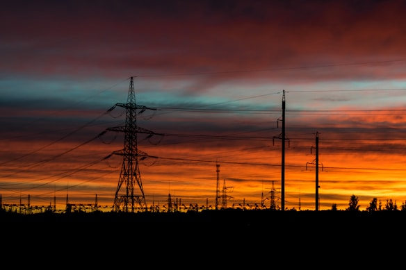 A series of electricity pylons and power lines are silhouetted against a vibrant sunset. The sky is painted with layers of orange, red, and blue hues, creating a dramatic backdrop. The landscape at the bottom shows trees and smaller structures in shadow, emphasizing the height and scale of the pylons.