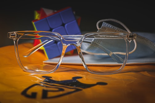 A pair of transparent eyeglasses positioned in the foreground, with a blue Rubik's cube and a blue surgical mask in the background. The items are placed on an orange surface with a black crown logo.