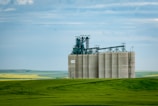A large industrial grain elevator stands prominently in a vast rural landscape, surrounded by lush green fields and under a partly cloudy blue sky. The structure is equipped with complex machinery and is a beige color, contrasting with the vibrant green and yellow fields in the background.