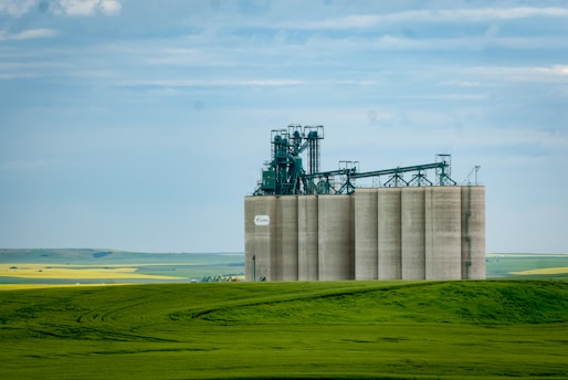 A large industrial grain elevator stands prominently in a vast rural landscape, surrounded by lush green fields and under a partly cloudy blue sky. The structure is equipped with complex machinery and is a beige color, contrasting with the vibrant green and yellow fields in the background.