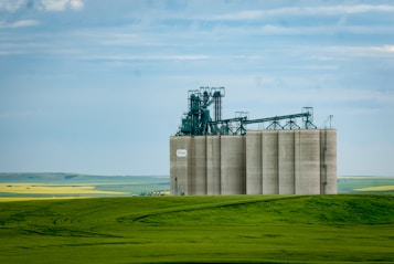 A large industrial grain elevator stands prominently in a vast rural landscape, surrounded by lush green fields and under a partly cloudy blue sky. The structure is equipped with complex machinery and is a beige color, contrasting with the vibrant green and yellow fields in the background.