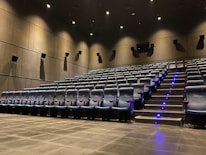 A modern cinema auditorium with rows of empty blue cushioned seats arranged in an ascending manner. The floor is covered with dark carpeting, and there are small blue lights along the stairway for safety. Wall-mounted speakers are visible, and the lighting is dim, focused mainly on illuminating the aisle.