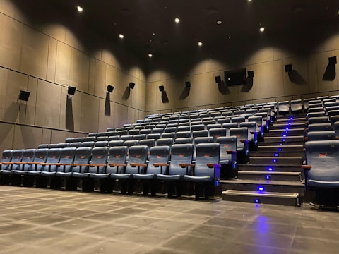A modern cinema auditorium with rows of empty blue cushioned seats arranged in an ascending manner. The floor is covered with dark carpeting, and there are small blue lights along the stairway for safety. Wall-mounted speakers are visible, and the lighting is dim, focused mainly on illuminating the aisle.