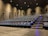 A modern cinema auditorium with rows of empty blue cushioned seats arranged in an ascending manner. The floor is covered with dark carpeting, and there are small blue lights along the stairway for safety. Wall-mounted speakers are visible, and the lighting is dim, focused mainly on illuminating the aisle.