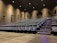 A modern cinema auditorium with rows of empty blue cushioned seats arranged in an ascending manner. The floor is covered with dark carpeting, and there are small blue lights along the stairway for safety. Wall-mounted speakers are visible, and the lighting is dim, focused mainly on illuminating the aisle.