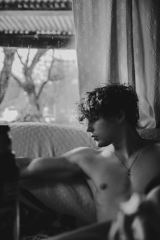 A young man with curly hair sits shirtless on a couch, gazing thoughtfully out of a window. The background features a curtain and a blurred view outside, with the silhouette of trees visible through the droplets on the glass. The image conveys a quiet, introspective moment captured in black and white.