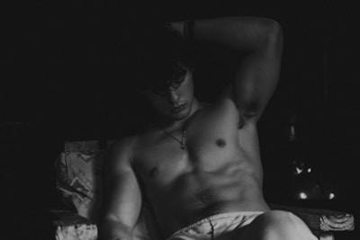 A shirtless person sits in a contemplative pose, with their arm raised and resting on their head. The scene is captured in black and white, emphasizing shadows and highlights on their muscular physique. The background is dark, adding a dramatic and moody atmosphere.