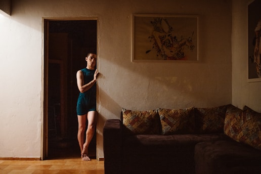 A person stands in a doorway with a thoughtful expression, wearing a blue outfit. The walls are adorned with framed artwork, and there is a dark-colored sofa with multi-patterned cushions. The lighting is warm and casts soft shadows, creating an intimate atmosphere.