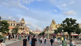 A bustling street in a theme park lined with early 20th-century style buildings. The street is filled with visitors enjoying their day, with many people walking, taking photos, and exploring. Tall trees and street lamps line the avenue, creating a welcoming atmosphere. The sky is partly cloudy with patches of blue.