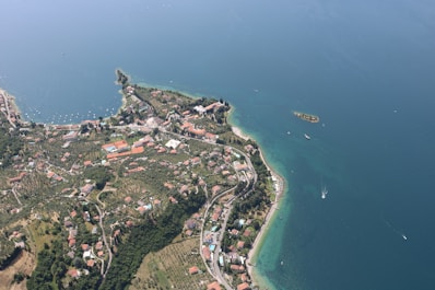 An aerial view captures a coastal area with sprawling greenery and dense clusters of buildings. The shoreline is defined by clear blue waters, and boats can be seen navigating through the sea. Roads meander through the landscape, highlighting a blend of natural and developed environments.