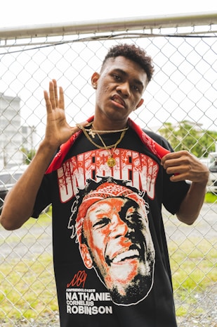 A person standing in front of a chain-link fence, wearing a graphic T-shirt and gold jewelry. The T-shirt features a large print of another person's face with the word 'UNFRIEND' above it and the name 'Nathaniel Cornelius Robinson' below. A red bandana is draped around the neck, and the person is posing with a hand gesture.