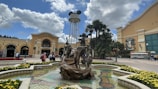 A lively theme park entrance with a large fountain in the foreground adorned with statues of characters. The background features an iconic water tower with mouse ears, surrounded by palm trees and colorful flowerbeds. Visitors are seen walking around, adding to the bustling atmosphere.