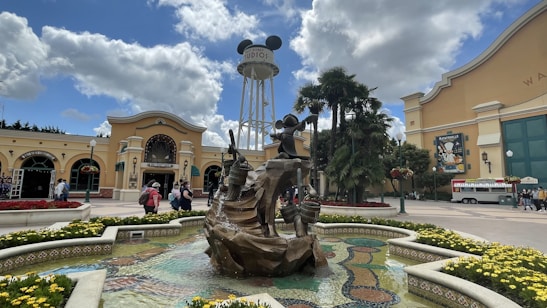 A lively theme park entrance with a large fountain in the foreground adorned with statues of characters. The background features an iconic water tower with mouse ears, surrounded by palm trees and colorful flowerbeds. Visitors are seen walking around, adding to the bustling atmosphere.