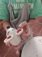 A person wearing a green shirt and a grey apron with white dots is holding a small, painted sculpture of a mouse with closed eyes and pink inner ears. The person's hands are covered in clay, indicating involvement in pottery or sculpting activity.