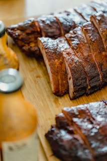 Slices of barbecue ribs are arranged on a wooden cutting board. The ribs have a rich, dark, glazed appearance, suggesting they are well seasoned and cooked. Bottles of sauces can be seen in the foreground, slightly out of focus.