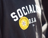 Text on a black garment says 'SOCIALISM D.S.A.' with a bright yellow circular sticker on it. The sticker features a flower design with the text 'DANDELION UNION ILWU LOCAL 6'.