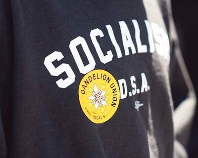 Text on a black garment says 'SOCIALISM D.S.A.' with a bright yellow circular sticker on it. The sticker features a flower design with the text 'DANDELION UNION ILWU LOCAL 6'.