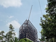 A large steel framework structure is under construction with a crane positioned at the top. Surrounding the structure are several tall green trees, and the sky is partly cloudy with a light blue hue.