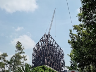 A large steel framework structure is under construction with a crane positioned at the top. Surrounding the structure are several tall green trees, and the sky is partly cloudy with a light blue hue.