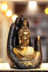A golden Buddha statue is nestled in a large, decorative black hand adorned with glitter. The background is softly illuminated by blurred warm lights, creating a serene and sacred atmosphere.