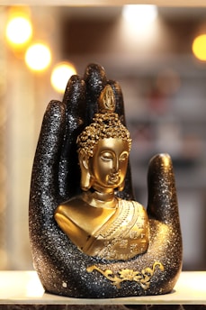 A golden Buddha statue is nestled in a large, decorative black hand adorned with glitter. The background is softly illuminated by blurred warm lights, creating a serene and sacred atmosphere.