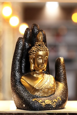 A golden Buddha statue is nestled in a large, decorative black hand adorned with glitter. The background is softly illuminated by blurred warm lights, creating a serene and sacred atmosphere.