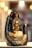 A golden Buddha statue is nestled in a large, decorative black hand adorned with glitter. The background is softly illuminated by blurred warm lights, creating a serene and sacred atmosphere.
