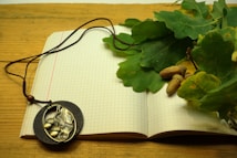 An open notebook with graph paper lies on a wooden surface. A few green oak leaves and acorns rest on top of the notebook, creating a natural autumn theme. A pendant with a leaf and acorn design connected to a thin brown cord is draped across the pages.