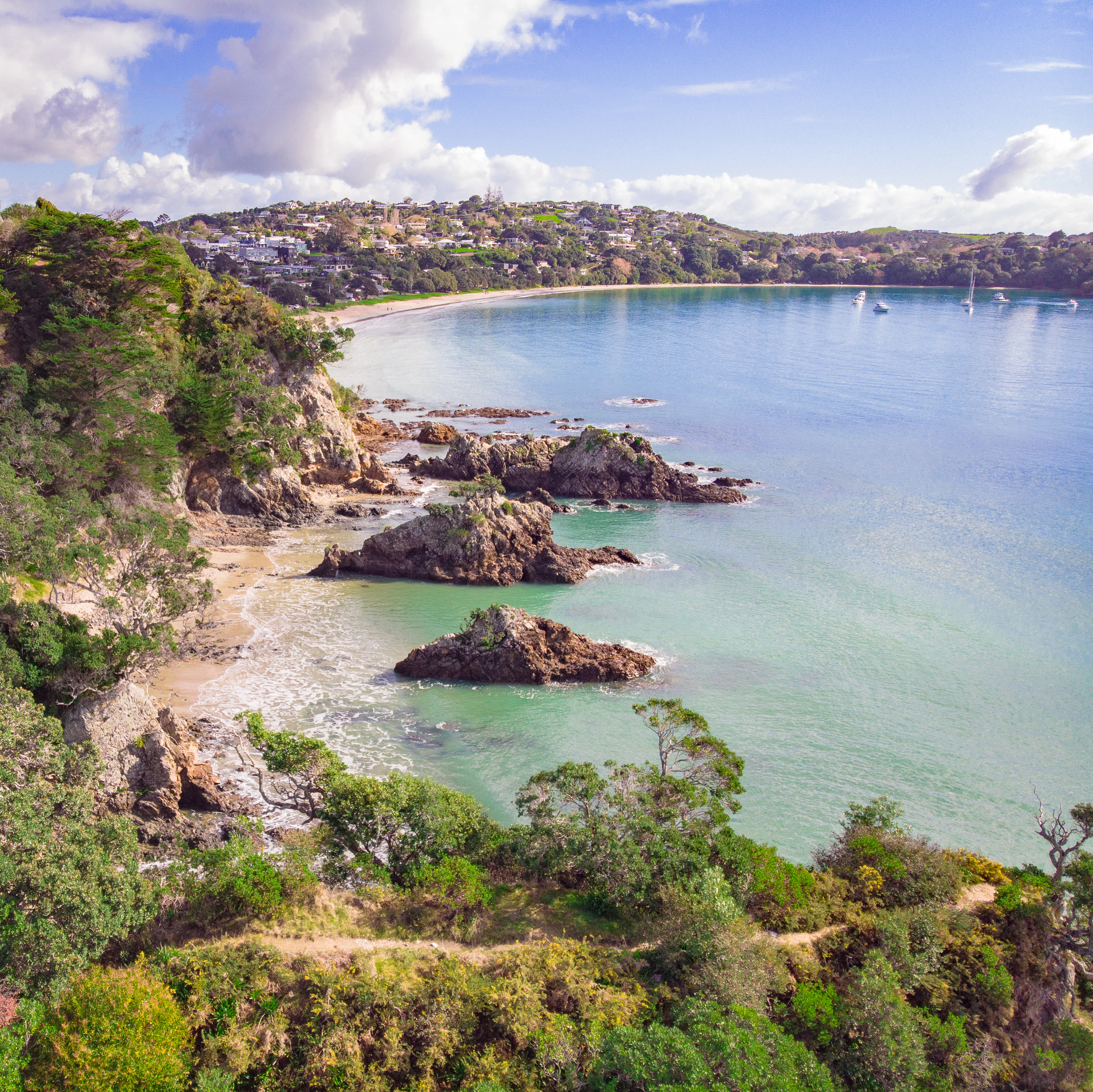 Aerial view of a turquoise Waiheke Island bay with a sandy beach, clifftop houses and moored boats.