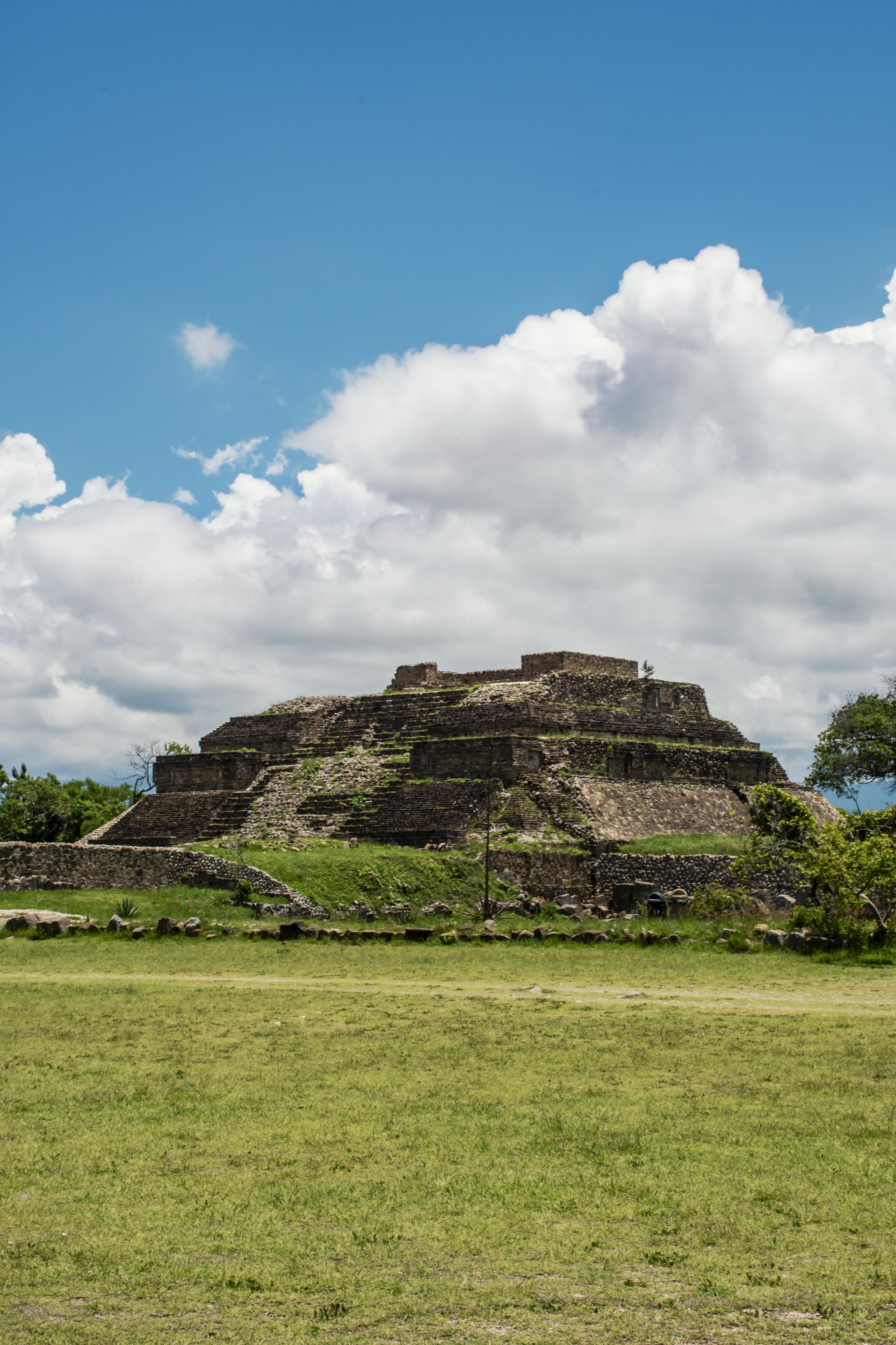 A stepped stone pyramid beyond a green plaza under billowing clouds at Monte Albán, Oaxaca