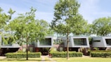 A modern residential building with large windows, surrounded by lush green trees and well-maintained shrubs. The structure features contemporary architecture with clean lines and a combination of white and brown colors. The sunny day enhances the vibrant greenery and the overall peaceful atmosphere of the location.