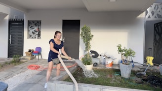 A woman is watering plants outside a modern house. She is holding a hose and standing on a tiled walkway. The house has a minimalist design with a black door, geometric pattern windows, and a covered porch. There are potted plants, scattered gardening tools, and a hose reel nearby.