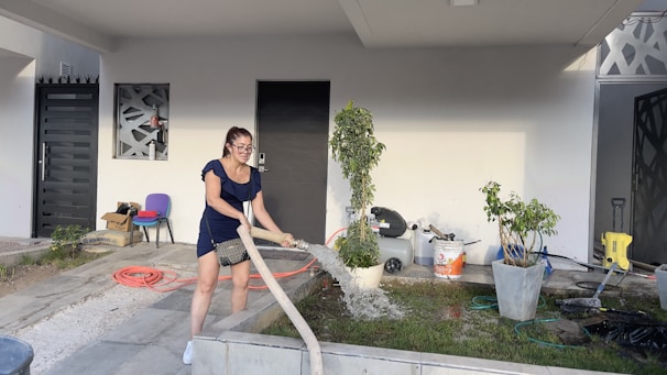 A woman is watering plants outside a modern house. She is holding a hose and standing on a tiled walkway. The house has a minimalist design with a black door, geometric pattern windows, and a covered porch. There are potted plants, scattered gardening tools, and a hose reel nearby.