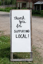 A sign with handwritten text saying 'thank you for supporting local!' placed outdoors near a rustic building. The background features greenery and a dirt path.