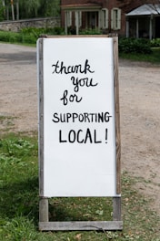 A sign with handwritten text saying 'thank you for supporting local!' placed outdoors near a rustic building. The background features greenery and a dirt path.