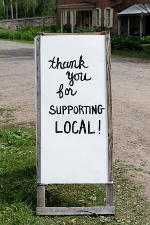 A sign with handwritten text saying 'thank you for supporting local!' placed outdoors near a rustic building. The background features greenery and a dirt path.