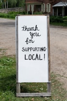 A sign with handwritten text saying 'thank you for supporting local!' placed outdoors near a rustic building. The background features greenery and a dirt path.