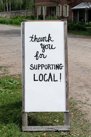 A sign with handwritten text saying 'thank you for supporting local!' placed outdoors near a rustic building. The background features greenery and a dirt path.
