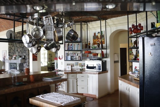 A spacious kitchen features hanging stainless steel pots, pans, and a grater from an overhead rack. Wooden countertops and white cabinetry line the walls, with a microwave placed on one counter near a collection of books and decorative vases on shelves. A black refrigerator stands to the right, and a kitchen island with a folded cloth is centered under the hanging cookware. The back wall has a selection of artwork and small appliances, adding to the cozy, lived-in atmosphere.
