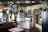 A spacious kitchen features hanging stainless steel pots, pans, and a grater from an overhead rack. Wooden countertops and white cabinetry line the walls, with a microwave placed on one counter near a collection of books and decorative vases on shelves. A black refrigerator stands to the right, and a kitchen island with a folded cloth is centered under the hanging cookware. The back wall has a selection of artwork and small appliances, adding to the cozy, lived-in atmosphere.