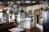 A spacious kitchen features hanging stainless steel pots, pans, and a grater from an overhead rack. Wooden countertops and white cabinetry line the walls, with a microwave placed on one counter near a collection of books and decorative vases on shelves. A black refrigerator stands to the right, and a kitchen island with a folded cloth is centered under the hanging cookware. The back wall has a selection of artwork and small appliances, adding to the cozy, lived-in atmosphere.