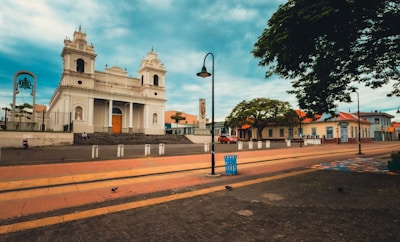 A large, historic church with a white facade and twin bell towers stands prominently in an open plaza. The surrounding area features paved walkways, street lamps, and a few scattered benches. There is a tree-lined street with a few vehicles parked alongside and people walking by. The sky above is partly cloudy, adding to the serene atmosphere.