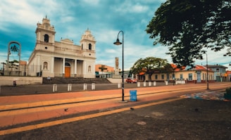 A large, historic church with a white facade and twin bell towers stands prominently in an open plaza. The surrounding area features paved walkways, street lamps, and a few scattered benches. There is a tree-lined street with a few vehicles parked alongside and people walking by. The sky above is partly cloudy, adding to the serene atmosphere.