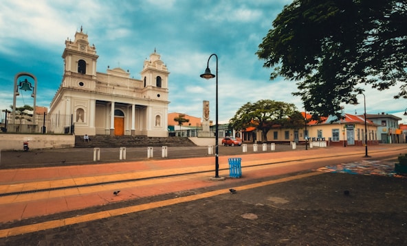 A large, historic church with a white facade and twin bell towers stands prominently in an open plaza. The surrounding area features paved walkways, street lamps, and a few scattered benches. There is a tree-lined street with a few vehicles parked alongside and people walking by. The sky above is partly cloudy, adding to the serene atmosphere.