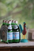 A pack of six sparkling water bottles with green glass and blue labels is placed on a surface. Beside it, there is a colorful rooster figurine and a lit candle in a glass container. The background shows a blurry, natural, green setting.