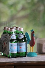 A pack of six sparkling water bottles with green glass and blue labels is placed on a surface. Beside it, there is a colorful rooster figurine and a lit candle in a glass container. The background shows a blurry, natural, green setting.