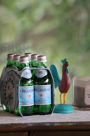 A pack of six sparkling water bottles with green glass and blue labels is placed on a surface. Beside it, there is a colorful rooster figurine and a lit candle in a glass container. The background shows a blurry, natural, green setting.