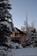 A wooden house with large windows is surrounded by snow-covered trees and shrubs. The roof has a sloped design, and the setting sun casts a warm glow on the house's facade. The sky is clear and blue, enhancing the tranquility of the winter scene.